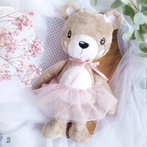 Puede incluir: Un oso de peluche marrón que lleva un tutú de tul rosa. El oso tiene la barriga blanca y los ojos negros.