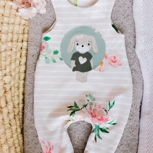 May include: White and green striped baby overalls with a floral print and a green and brown applique of a bunny with a heart on its shirt.
