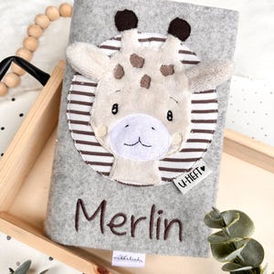 May include: A gray felt book cover featuring a giraffe design with brown spots and a white face. The name "Merlin" is embroidered in brown. A small tag reads "U-HEFT".