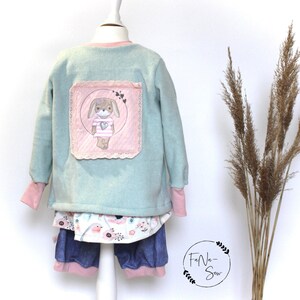 May include: A light blue fleece jacket with a pink patch featuring a cartoon bunny with a heart. The jacket has long sleeves and a pink trim around the neck.