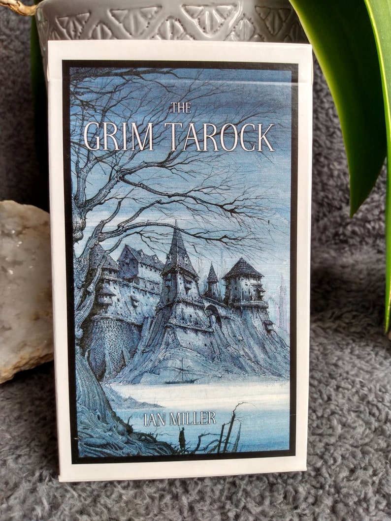 以下が含まれることがあります： 「The Grim Taroc」というタロットカードデッキの箱。暗いゴシック様式の城が雪景色の中に描かれている。アーティストの名前「Ian Miller」がタイトルの下に印刷されている。