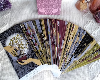 Puede incluir: Una baraja de cartas de tarot desplegada en semicírculo, mostrando el dorso de las cartas. Las cartas tienen un fondo oscuro con una variedad de colores y patrones. La carta en la parte delantera de la baraja muestra una mano sosteniendo una pequeña planta en una maceta. El texto en la carta dice "O Meigo..."