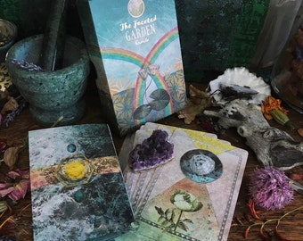 May include: A box of The Faceted Garden Oracle cards, with a colorful illustration of a rainbow and lotus flowers. The cards are spread out on a wooden table with a crystal, flowers, and other decorative items.