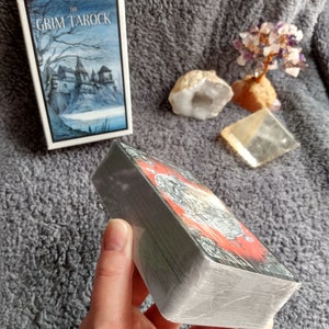 以下が含まれることがあります： グリム・タロック・タロットカードのデッキ。暗いファンタジーをテーマにしたデッキで、箱には城の白黒のイラストが描かれています。カードは積み重ねられ、手で持たれています。
