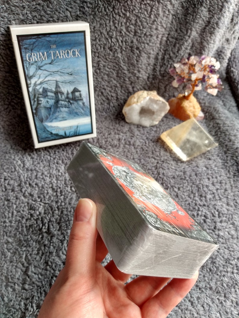 以下が含まれることがあります： グリム・タロック・タロットカードのデッキ。暗いファンタジーをテーマにしたデッキで、箱には城の白黒のイラストが描かれています。カードは積み重ねられ、手で持たれています。