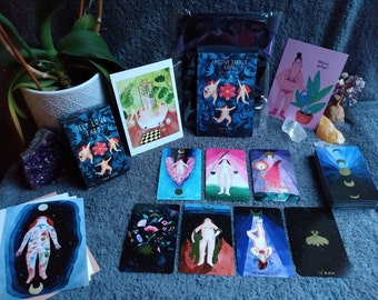 May include: A set of tarot cards with colorful illustrations on a blue background. The cards are laid out on a gray surface with other items, including a book titled "Positive Energy Guide", a card with the text "Grow Bitch", and a small potted plant.