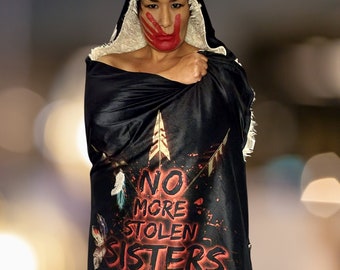 May include: A person wearing a black hooded blanket with the words "NO MORE STOLEN SISTERS" printed in red. The person has red paint on their face and hands.