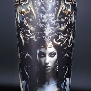 May include: A stainless steel tumbler with a black and gold design featuring Medusa, a Greek mythological figure with snakes for hair. The design includes a Greek key border.