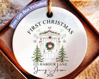 Puede incluir: Un adorno redondo de cerámica blanca con cuerda de yute, que presenta una ilustración de una puerta principal con corona, dos pinos y el texto FIRST CHRISTMAS AT 123 HABOUR LANE, Jenny + Anson, y el año 2026.