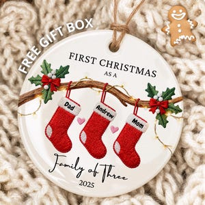 Op de afbeelding: Een ronde witte keramische kerstversiering met een familiethema. Het heeft drie rode kousen die aan een tak hangen, elk met een naam. De tekst luidt "First Christmas as a Family of Three 2025". Een peperkoekmannetje staat op de achtergrond.