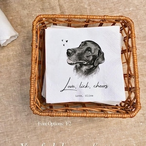 May include: A stack of white paper napkins inside a square woven wicker basket. The top napkin features a black and white sketch of a dog, three small hearts, and the text "Love, lick, cheers" followed by "Love, Olive" in black ink.