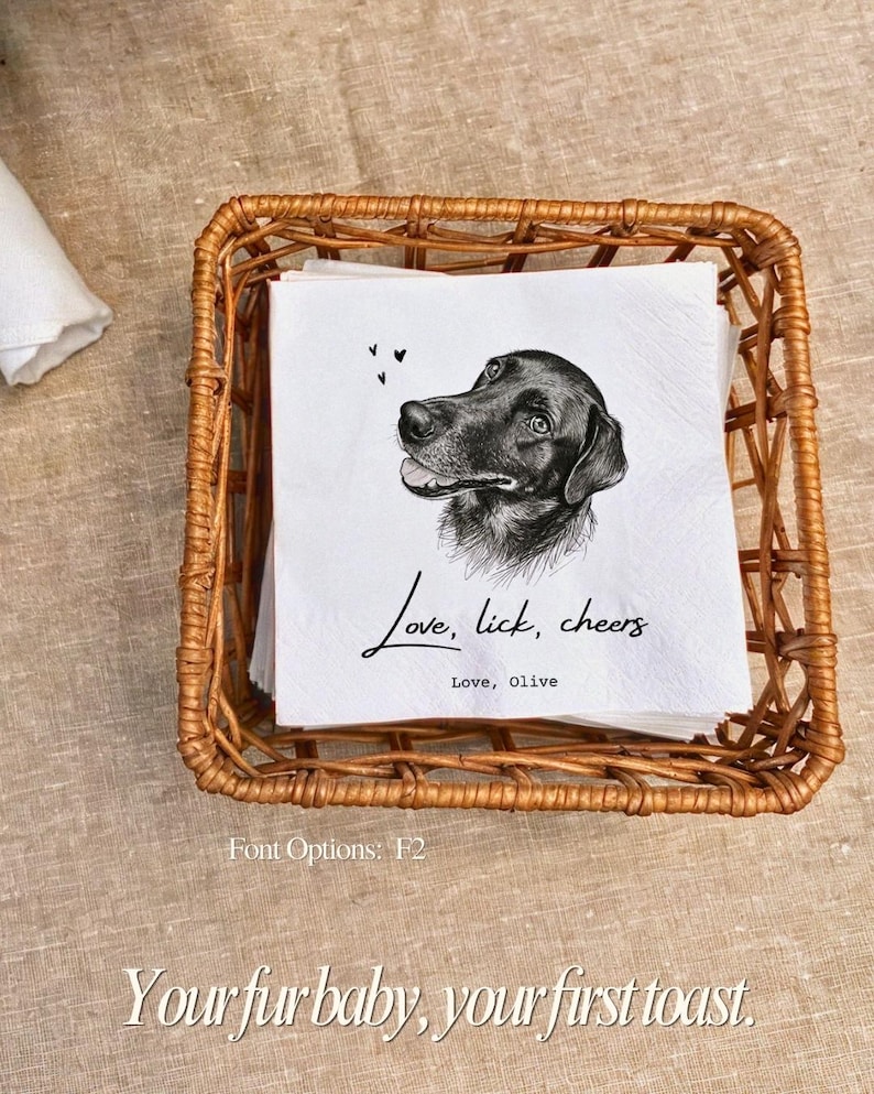 May include: A stack of white paper napkins inside a square woven wicker basket. The top napkin features a black and white sketch of a dog, three small hearts, and the text "Love, lick, cheers" followed by "Love, Olive" in black ink.