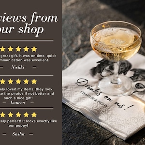 May include: A white paper napkin featuring a black and white illustration of two dogs, with the text "Drinks on us!" and "CHARLIE & PIGGY" printed in black. A clear glass filled with a golden liquid sits on top of the napkin.