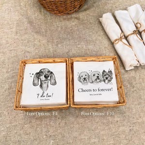May include: Two square wicker baskets containing stacks of white paper napkins. The left stack features a dachshund illustration with the text "I do too! Love, Buster" and "Font Options: F4". The right stack shows three dogs with "Cheers to forever! Sera, Leo & Alfie" and "Font Options: F10".