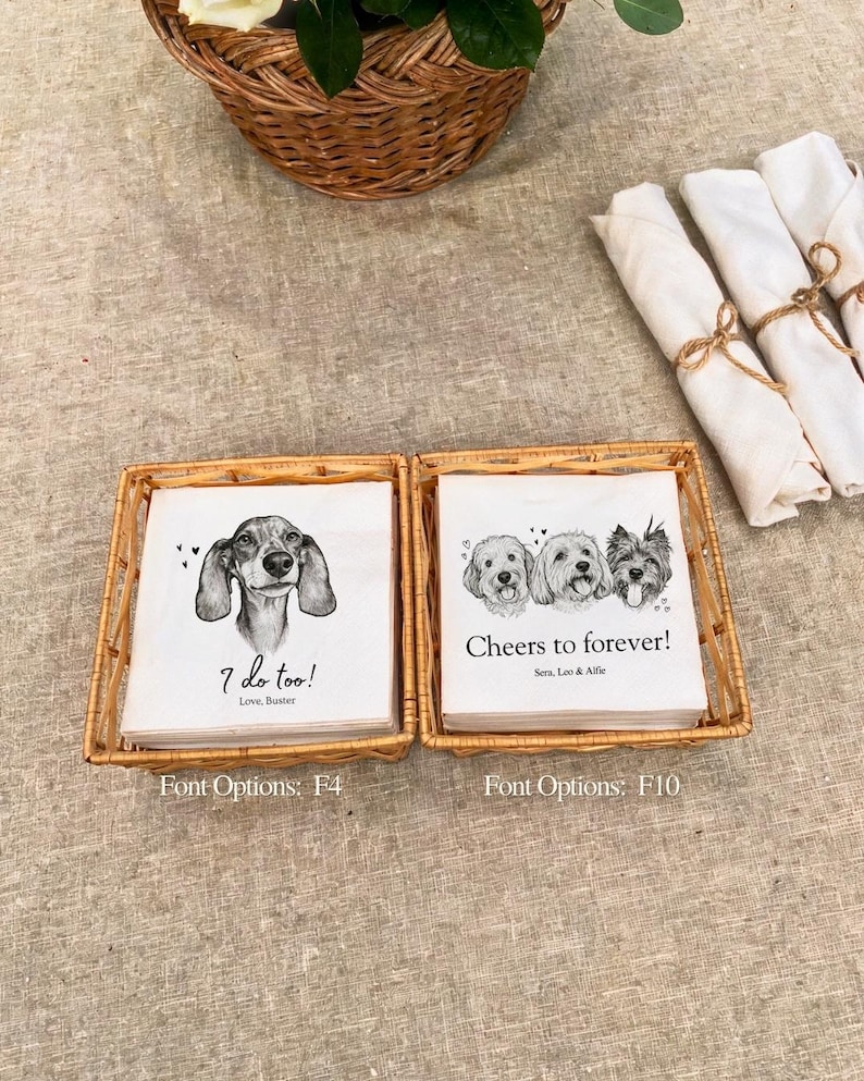 May include: Two square wicker baskets containing stacks of white paper napkins. The left stack features a dachshund illustration with the text "I do too! Love, Buster" and "Font Options: F4". The right stack shows three dogs with "Cheers to forever! Sera, Leo & Alfie" and "Font Options: F10".