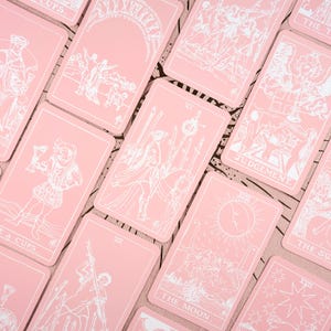 May include: A collection of pink tarot cards with white line art illustrations. The cards feature various figures, symbols, and text, including "Judgement" and "The Moon". The cards are arranged in a scattered pattern.