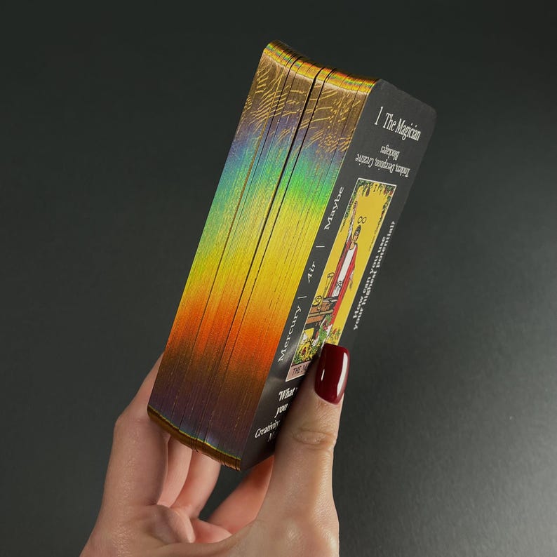 May include: A deck of tarot cards with iridescent edges, held in a hand. The card faces feature colorful illustrations and text, including "The Magician" and "Mercury | Air | Maybe". The edges of the cards have a rainbow effect.