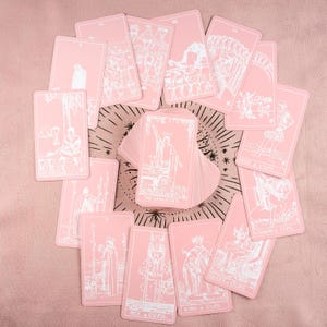 May include: A deck of pink tarot cards with white illustrations, including "The Magician," "Ace of Cups," and "Queen of Cups." The cards are arranged in a circular pattern on a pink surface, with some cards fanned out. The deck is a divination tool.