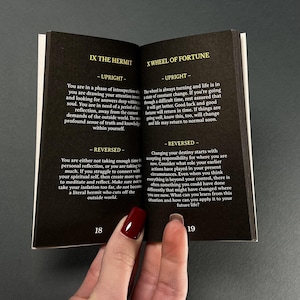 May include: An open book with black pages, featuring text about tarot card meanings. The left page displays "IX THE HERMIT" and the right page shows "X WHEEL OF FORTUNE". The pages are held open by a hand with red nail polish.