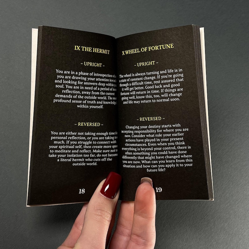 May include: An open book with black pages, featuring text about tarot card meanings. The left page displays "IX THE HERMIT" and the right page shows "X WHEEL OF FORTUNE". The pages are held open by a hand with red nail polish.