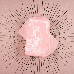 May include: A deck of pink tarot cards fanned out on a pink surface with a sunburst design. The top card features a white illustration of a figure with the text "THE MAGICIAN". The cards are a soft pink color.