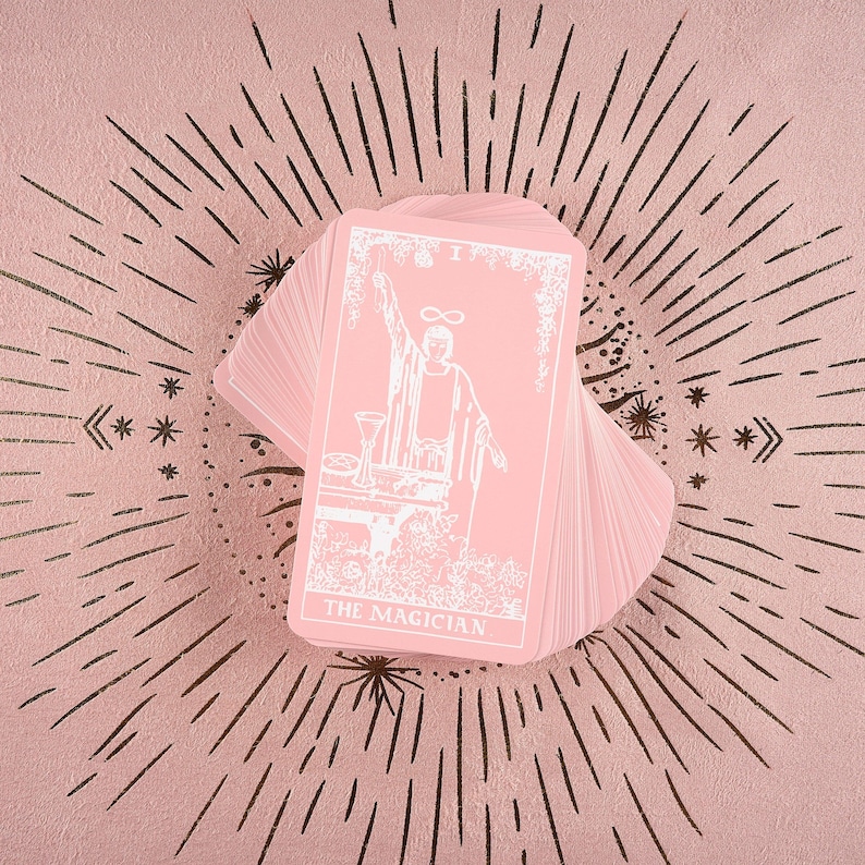 May include: A deck of pink tarot cards fanned out on a pink surface with a sunburst design. The top card features a white illustration of a figure with the text "THE MAGICIAN". The cards are a soft pink color.