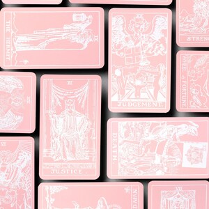 May include: A collection of pink tarot cards with white illustrations and text. The cards feature various designs, including figures, symbols, and the words "JUDGEMENT" and "JUSTICE". The cards are arranged on a black surface.