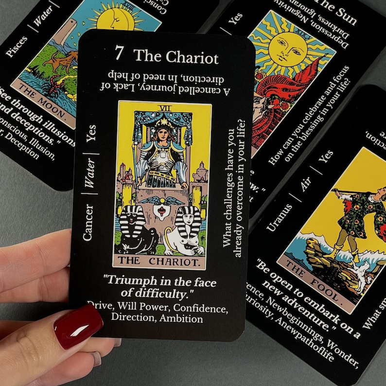 May include: A collection of black tarot cards with gold and white illustrations. The central card, "The Chariot," features a figure in a chariot. Other cards include "The Moon," "The Sun," and "The Fool." Text on the cards includes keywords and phrases.