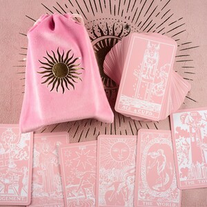 May include: A pink tarot card deck with white illustrations and text, including "ACE 4 CUPS" and "THE WORLD". The deck is accompanied by a pink velvet drawstring bag with a gold sun design. The cards are spread out on a pink surface.