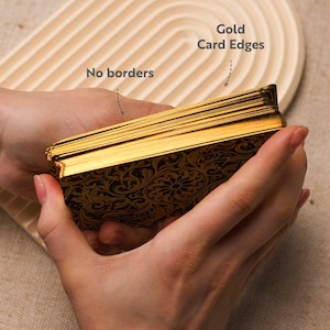 May include: A deck of tarot cards with gold edges and a black and gold floral pattern. The cards are held in two hands. The text "Gold Card Edges" and "No borders" are visible.