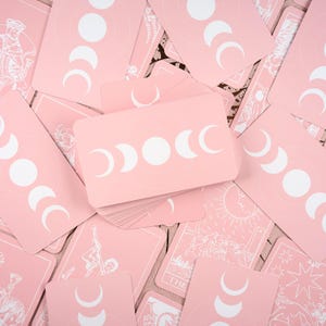 May include: A collection of pink tarot cards with white celestial designs. The backs of the cards feature a crescent moon phase design. Some cards show illustrations of the sun, stars, and figures. The cards are scattered on a wooden surface.