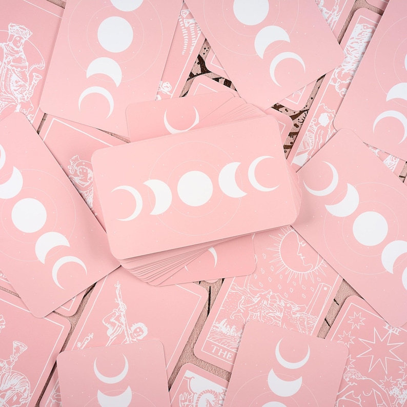 May include: A collection of pink tarot cards with white celestial designs. The backs of the cards feature a crescent moon phase design. Some cards show illustrations of the sun, stars, and figures. The cards are scattered on a wooden surface.