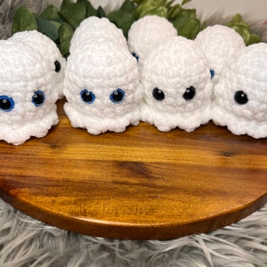 May include: Five crocheted ghost ornaments with blue eyes. The ghosts are white and have a textured surface. They are arranged on a wooden surface.