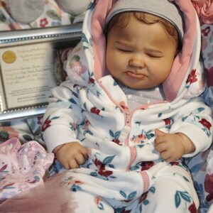 May include: A lifelike baby doll wearing a white onesie with a floral pattern of red and blue flowers. The doll has a gray beanie and a hooded jacket with a pink interior. The doll is lying on a floral blanket.