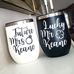 Könnte beinhalten: Zwei weingläser ohne Stiel, eines weiß und eines schwarz, mit silbernen Rändern. Auf dem weißen Becher steht "Future Mrs Keane" mit einer Ringgrafik. Auf dem schwarzen Becher steht "Lucky Mr Keane" mit einem vierblättrigen Kleeblatt.