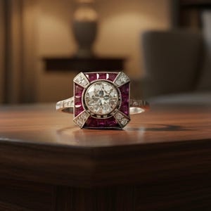 Peut inclure: Une bague de style vintage avec une grosse pierre transparente ronde au centre, entourée de petites pierres rouges et transparentes. La bague est posée sur une surface en bois sombre, avec une lampe et un arrière-plan flou.