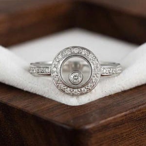 May include: A silver ring with a floating diamond center and a halo of smaller diamonds. The ring is set on a white velvet surface inside a dark wooden box, showcasing the ring's sparkle and design.