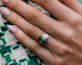 May include: A gold ring featuring a large emerald-cut green gemstone and a pear-shaped clear gemstone. The ring is set on a hand with manicured nails, against a green and white patterned background.