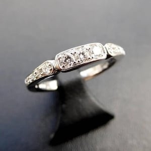 May include: A silver ring with a row of five clear gemstones set in a rectangular design. The ring has smaller gemstones on the sides and is displayed on a black stand against a dark background.