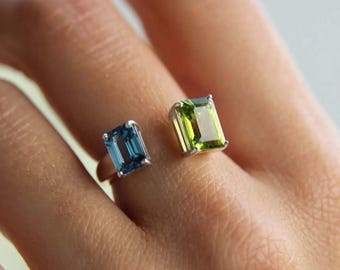 May include: Two rings with rectangular gemstones are displayed. One ring features a blue gemstone, the other a green gemstone. Both gemstones are set in silver bands. The rings are shown on a person's finger.