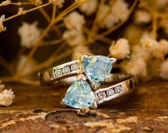 May include: A silver ring with two triangular blue gemstones set in the band. The ring is on a brown wooden surface with dried flowers in the background.