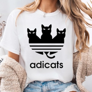 May include: White t-shirt with a black graphic of three black cats in a triangle formation with a tail extending down to the text "adicats".