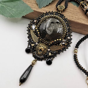Peut inclure: Un collier de perles noires et dorées avec un grand pendentif représentant une tête de lion, des ailes et une pierre noire. Le pendentif est entouré de perles noires et dorées.