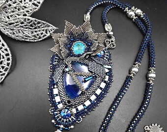 Puede incluir: Un collar de cuentas azul y plateado con un gran colgante intrincado que presenta una piedra de ágata azul y cuentas azules y plateadas. El colgante tiene la forma de una flor con un centro puntiagudo.