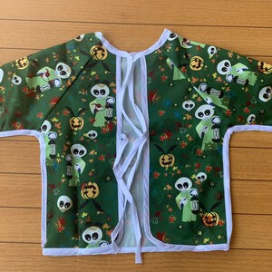 May include: Green and white long-sleeved smock with a tie closure at the back. The smock has a Halloween theme with a repeating pattern of green ghosts holding lanterns and orange pumpkins.
