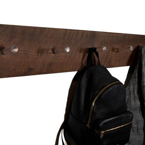 May include: A dark brown wooden wall mounted coat rack with five clear acrylic hooks. A black leather backpack and a gray jacket are hanging on the hooks.