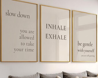 May include: Three vertical wall art prints in thin gold frames featuring beige backgrounds and dark gray text. The first print reads slow down you are allowed to take your time. The second reads INHALE EXHALE. The third reads be gentle with yourself you are still growing.