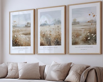 May include: Three framed landscape art prints with a neutral color palette. Each print features a field of wildflowers with text from the Bible. The frames are a light wood color, and the prints are displayed above a beige sofa.