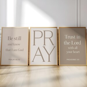 Può includere: Tre stampe da parete rettangolari in sottili cornici dorate. La stampa a sinistra presenta il testo Be still and know that I am God PSALM 46:10. La stampa centrale mostra la parola PRAY. La stampa a destra mostra Trust in the Lord with all your heart PROVERBS 3:5.
