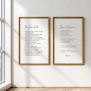 May include: Two framed prints with gold-colored frames hang on a white wall. The prints feature black text on a white background. The left print is titled "Du Zahn Gebote" and the right print is titled "Das Vaterunser". The prints are displayed near a window.